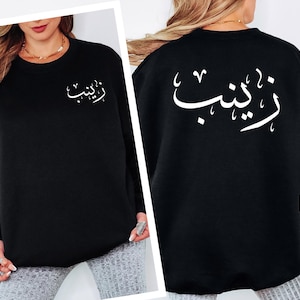 May include: Black sweatshirt with white Arabic script on the front and back. The script appears to be a name. The sweatshirt is a crew neck style and has long sleeves. The person is wearing a gold necklace.