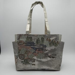 May include: A silver tote bag with a landscape design featuring a pagoda, trees, and mountains. The bag has a metallic silver handle and a pocket on each side. The landscape design includes shades of green, brown, and pink.