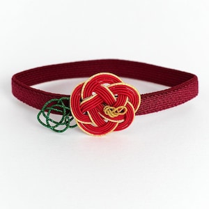 May include: A dark red elastic band featuring a decorative knot ornament made of red and gold cord. The ornament is shaped like a flower with a small green cord accent attached to the side, creating a detailed and textured accessory piece.