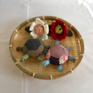 May include: Two handmade fabric turtle plushies with patchwork shells, green heads and limbs, and blue accents, sit in a woven bamboo basket. Two fabric flowers, one red and one white, are also in the basket. The basket is on a white surface.