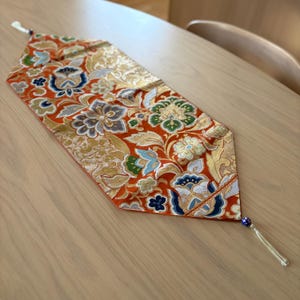 May include: An orange table runner with a floral pattern in gold, blue, green, and gray. The runner has a tassel at one end and is placed on a wooden table. The runner is approximately 12 inches wide.