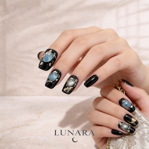 May include: Black nail polish with a design featuring a blue and silver orb with gold accents. The nails are square-shaped. The brand name "LUNARA" is at the bottom of the image.