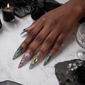 Puede incluir: Una mano con uñas largas, puntiagudas y decoradas. Las uñas presentan diseños que incluyen una serpiente, una cruz, un ojo y purpurina. La paleta de colores incluye verde, morado, plateado y rojo. Una vela negra, una rosa y otros elementos de temática gótica están en el fondo.