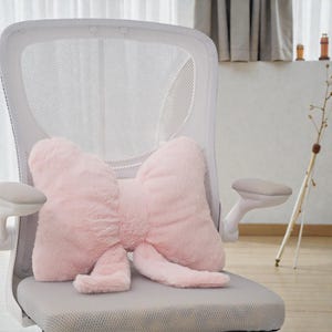 Puede incluir: Un cojín rosa claro en forma de lazo descansa sobre una silla de oficina de malla gris. El cojín tiene una apariencia esponjosa y texturizada, con un nudo central y extremos fluidos.