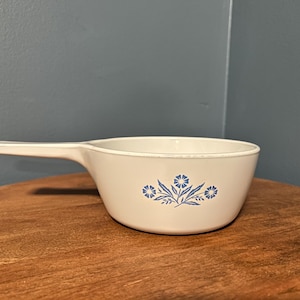 Puede incluir: Cacerola de cerámica blanquecina con un solo asa. La cacerola presenta un diseño floral azul en el lateral. La cacerola está sobre una superficie de madera.