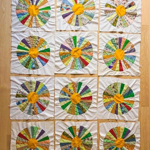 May include: Twelve individual quilt blocks featuring a colorful, circular design reminiscent of a flower. Each block has a yellow center and radiating segments of various patterned fabrics on a white background. The blocks are arranged on a wooden surface.