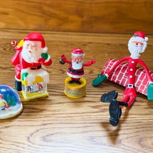 May include: A collection of Christmas-themed figurines, including a snow globe, a bendable Santa Claus, and two Santa Claus figures. The figurines are in red, white, and yellow. The snow globe features a winter scene.