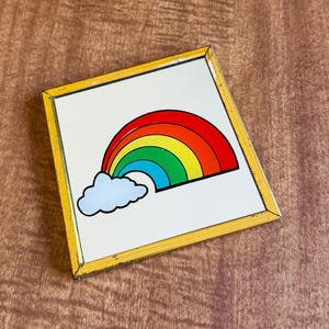 May include: A square, framed artwork featuring a colorful rainbow and a white cloud against a white background. The rainbow arcs across the top, displaying red, orange, yellow, green, and blue hues. The frame is yellow.
