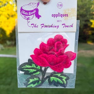 May include: A red embroidered rose applique with green and black leaves, packaged on a white card. The card reads "Cloth World" and "The Finishing Touch". The applique is in a clear plastic sleeve.