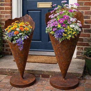 Könnte beinhalten: Zwei kegelförmige, rostige Metallpflanzgefäße gefüllt mit bunten Blumen. Eines hat orange, gelbe und violette Blumen, das andere violette, rosa, weiße und gelbe Blüten. Die Pflanzgefäße stehen auf runden Sockeln vor einer blauen Tür.