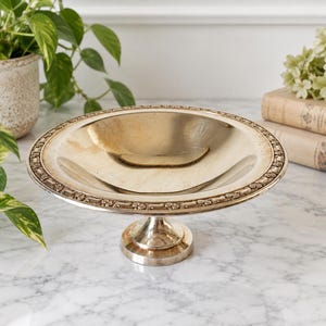 May include: A silver-toned pedestal bowl featuring a shallow, circular basin and a raised rim decorated with a repeating floral pattern. The bowl is supported by a central, flared pedestal base that rests on a circular foot, with a polished finish.