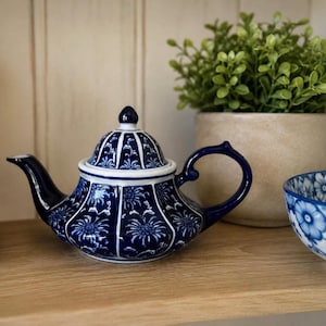 May include: A blue and white porcelain teapot with a floral design. The teapot has a curved spout, a rounded body, and a loop handle. The lid is topped with a small knob. A matching teacup is visible on the right.