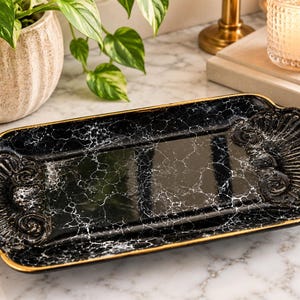 May include: A rectangular black tray with a white marble-like veined pattern and a glossy finish. The tray features ornate, shell-shaped decorative ends with raised scrollwork details and a thin gold-painted trim along the outer edges.