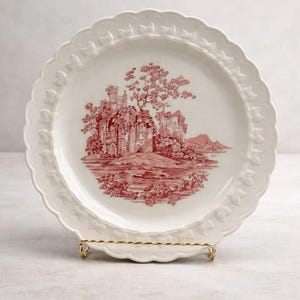 May include: White ceramic plate with a red castle scene. The plate has a scalloped edge with leaf detailing. The plate is displayed on a gold-colored metal stand. The castle scene includes trees, water, and mountains.