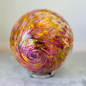 May include: A decorative glass sphere with a swirl design. The sphere features a blend of colors, including pink, yellow, and orange, with a clear base. The glass has a speckled pattern and a swirling design in the center.