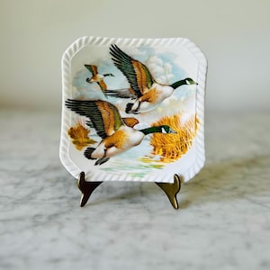 May include: A rectangular white decorative plate with a rope-like border. The plate features a detailed illustration of three geese in flight over a marshy landscape with yellow reeds and water. The plate is displayed on a gold-colored stand.