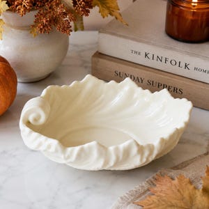 May include: A cream-colored ceramic bowl shaped like a large, curled leaf with a smooth, glossy finish. The bowl features intricate, embossed vein details radiating from the center toward the scalloped, wavy edges, creating a naturalistic form.