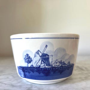 May include: White ceramic bowl with a blue and white windmill and landscape design. The bowl features a blue rim and a detailed illustration of a Dutch windmill, trees, and houses. The design is reminiscent of Delftware.