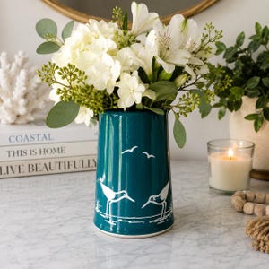 May include: A teal ceramic vase featuring a white line art illustration of two shorebirds and flying seagulls. The vase is filled with a bouquet of white flowers, including hydrangeas and alstroemeria, accented with green eucalyptus leaves.