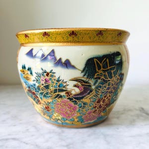 May include: A round ceramic planter featuring a cream-colored base with a hand-painted landscape scene. The design includes purple mountains, vibrant pink and blue flowers, and a bird, all outlined in gold. The rim has a yellow patterned border.