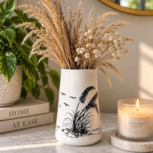 May include: A white ceramic vase featuring a black ink-style illustration of tall grasses and birds in flight. The vase is filled with dried wheat stalks and small white flowers, creating a natural arrangement with a clean, tapered silhouette.
