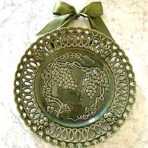 May include: A green ceramic decorative plate with a grape design. The plate has a pierced edge and is adorned with a green satin ribbon for hanging. The plate's diameter is approximately 10 inches.