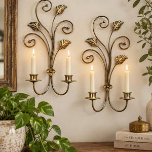 May include: Two matching wall-mounted candle sconces made of dark bronze-toned metal. Each sconce features a central floral accent, scrolling vine-like arms, and decorative leaves, with two candle holders each containing a lit white taper candle.
