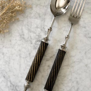 May include: A silver spoon and fork set with dark brown and gold striped handles. The silverware has a decorative design at the base of the handle and where the handle meets the spoon and fork heads. The set is displayed on a marble surface.