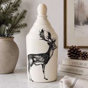 May include: A cream-colored ceramic canister with a tall, tapered neck and a rounded wooden stopper. The body features a detailed black and white illustration of a spotted deer with large antlers standing in profile on the front surface.