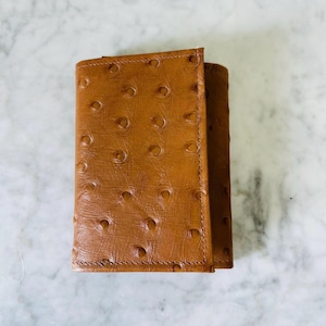 May include: A rectangular tri-fold wallet in a medium brown color featuring a textured surface with a repeating pattern of small, circular indentations across the exterior. The wallet has visible stitching along the edges and a folded flap closure.