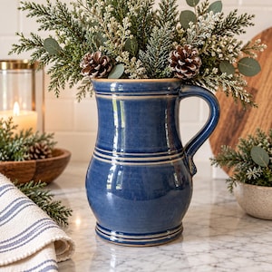 May include: A glossy, deep blue ceramic pitcher with a rounded body, a flared rim, and a single handle. The pitcher features horizontal ridged bands around the neck and base, with a smooth, reflective finish throughout the exterior surface.