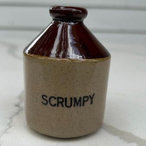 May include: A small, brown ceramic bottle with a dark brown top and the word "SCRUMPY" printed in black. The bottle has a cylindrical shape and a glossy finish on the top.