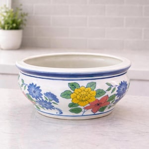 May include: A white ceramic bowl with a blue rim and floral designs. The bowl features a variety of colorful flowers, including yellow, red, and blue, with green leaves. The bowl is likely used for decorative purposes or as a planter.