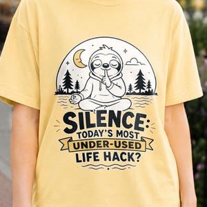 May include: A butter yellow t-shirt featuring a graphic of a sloth sitting in a meditative pose with a finger to its lips. The text reads: SILENCE: TODAY'S MOST UNDER-USED LIFE HACK? The design includes a crescent moon, stars, and pine trees.