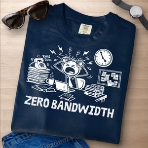 Puede incluir: Camiseta azul marino con un gráfico blanco de una persona estresada en un escritorio, rodeada de un teléfono sonando, un portátil y un reloj. El texto "ZERO BANDWIDTH" está impreso debajo. Gafas de sol y un reloj de pulsera también son visibles.