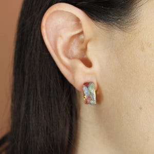 May include: A gold-coloured rectangular earring with a colourful, textured stone. The stone features shades of red, green, and blue. The earring is worn on an ear, with the person's hair and skin visible.