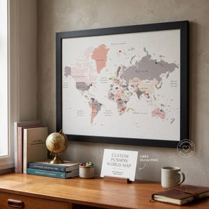 May include: A framed world map with push pins, in shades of pink, grey, and beige. The map is displayed above a wooden desk with books, a globe, and a mug. The text "CUSTOM PUSHPIN WORLD MAP" is visible on a card.