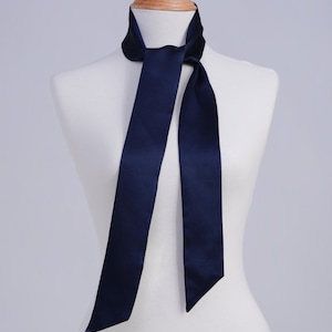 May include: A navy blue satin scarf is draped over a mannequin. The scarf is tied in a simple knot, with the long ends hanging down. The fabric has a subtle sheen, and the color is a deep, rich blue. The scarf is a classic accessory.