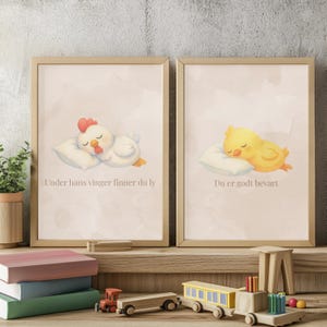 May include: Two framed wall art prints in light wood frames. The left print shows a white chicken sleeping on a pillow with the text Under hans vinger finner du ly. The right print shows a yellow chick sleeping on a pillow with the text Du er godt bevart.