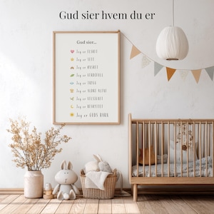 May include: A framed print with the text "Gud sier hvem du er" and a list of affirmations in Norwegian. The print is in a light wood frame and hangs on a white wall. The room is decorated with a crib, a basket of toys, and a decorative garland.