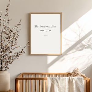 May include: A framed print with the text "The Lord watches over you" in a light-colored font. The print is in a light wood frame. The print is displayed above a wooden crib with a beige blanket and a stuffed teddy bear.