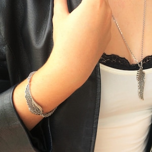 Puede incluir: Un collar y una pulsera de plata con un dise&ntilde;o de ala. El collar lo lleva una persona que lleva una chaqueta de cuero negra y un top blanco sin mangas.