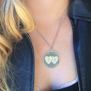 May include: Silver-toned pendant necklace with a round charm. The charm features two heart shapes and the engraved text "I AM YOURS YOU ARE MINE" with the initials "S" and "J". The necklace is worn against a black top and leather jacket.