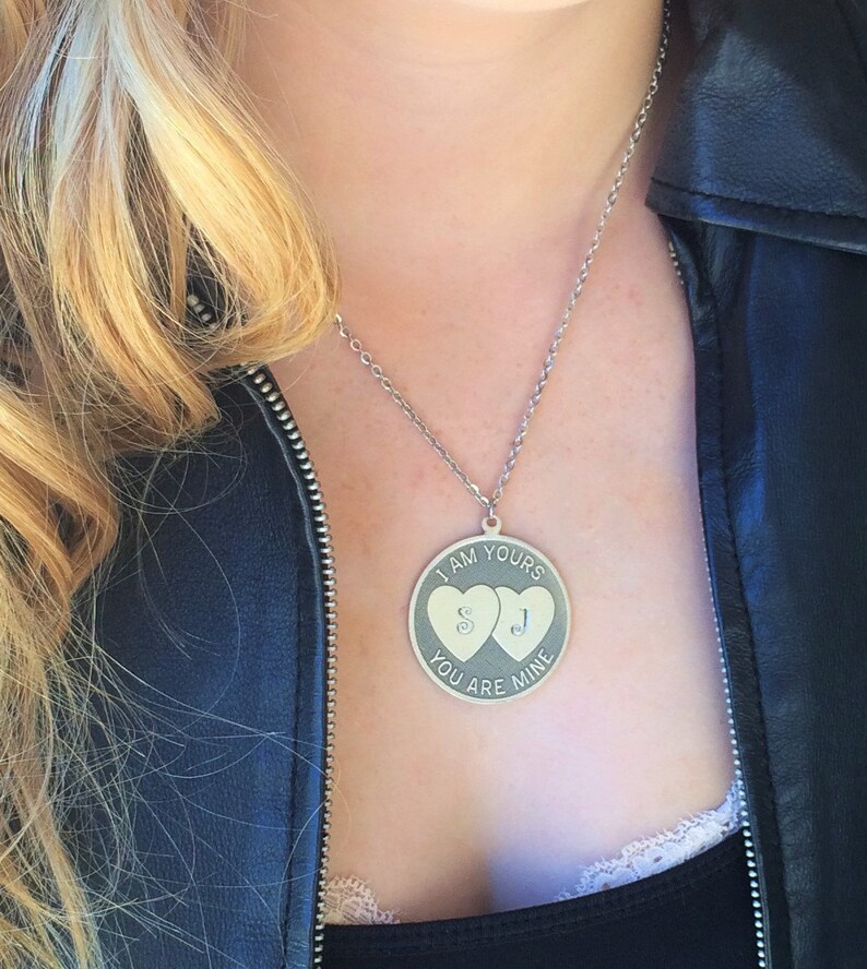 May include: Silver-toned pendant necklace with a round charm. The charm features two heart shapes and the engraved text "I AM YOURS YOU ARE MINE" with the initials "S" and "J". The necklace is worn against a black top and leather jacket.
