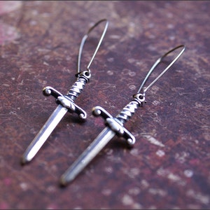 May include: A pair of silver dangle earrings featuring a sword design. The swords have a detailed, ornate design and are suspended from thin silver wires.