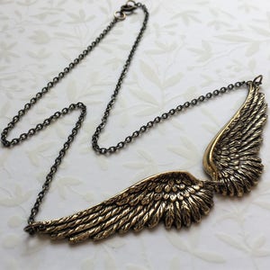 May include: A gold-toned necklace with a pair of angel wings pendant. The wings are detailed with a textured, feathery design. The necklace is made of a black chain.