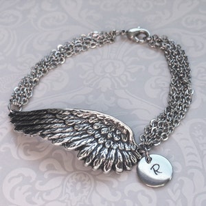 Puede incluir: Pulsera de plata con un dije de ala de &aacute;ngel detallado y un dije redondo con la letra 'R' grabada.
