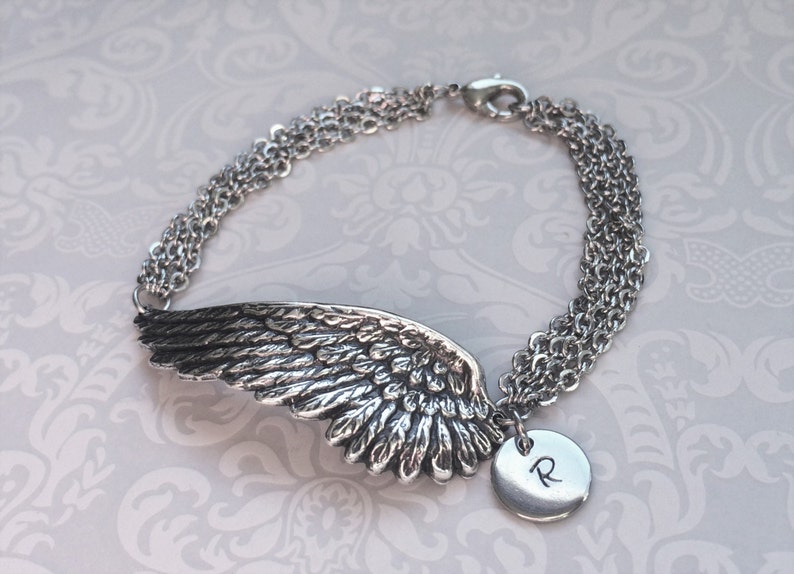 Puede incluir: Pulsera de plata con un dije de ala de &aacute;ngel detallado y un dije redondo con la letra 'R' grabada.