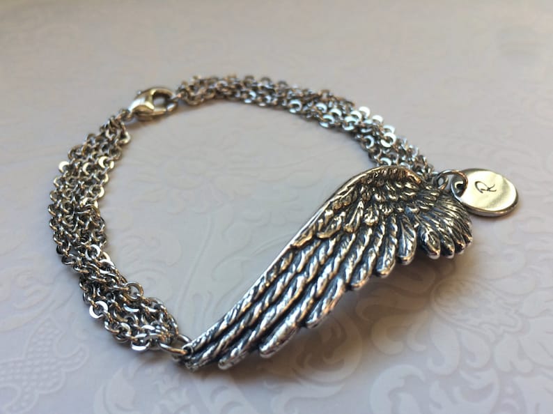 Puede incluir: Pulsera de plata con un dije de ala detallada y un dije redondo con la letra "R" grabada.