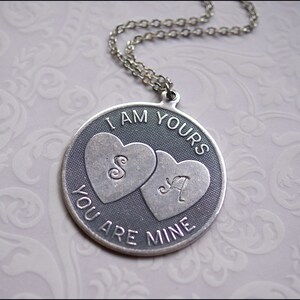 May include: A silver-toned pendant necklace with a circular design. The pendant features two heart shapes with the letters "S" and "A" inside. The words "I AM YOURS" and "YOU ARE MINE" are inscribed around the hearts. The necklace has a delicate chain.